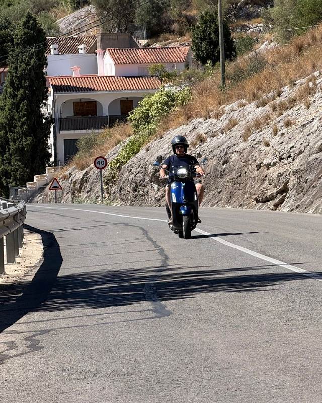 A rider on a blue Vespa coming down a mountain road past a village