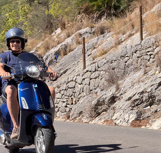 A rider on a blue Vespa Primavera beside a dry-stone wall
