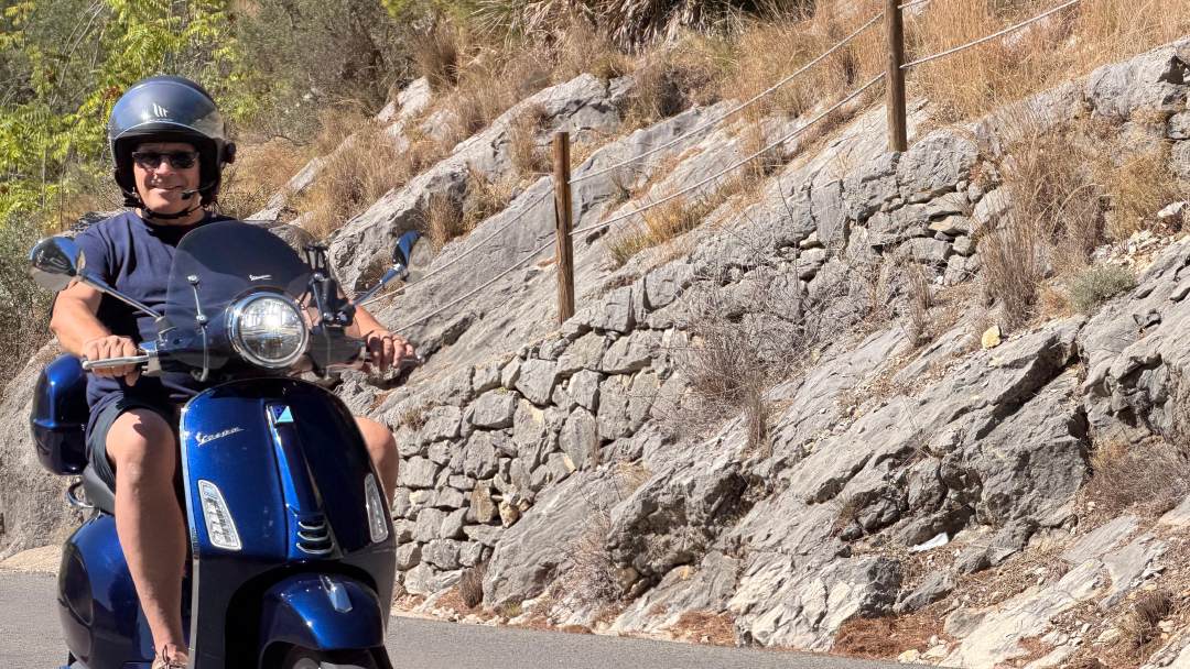 A rider on a blue Vespa Primavera on a quiet mountain road